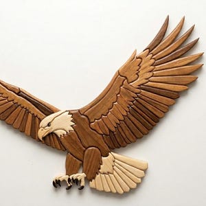 Puede incluir: Escultura de águila de madera con las alas extendidas, elaborada con varias tonalidades de madera marrón y beige. La cabeza y las plumas de la cola del águila son de color más claro, creando contraste. La escultura está montada sobre un fondo blanco.