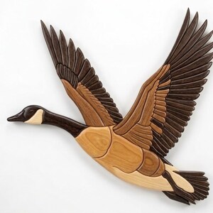 May include: Wooden wall art of a Canada goose in flight. The goose is crafted from various shades of wood, including light brown, dark brown, and black. The wings are spread wide, showcasing the detailed feather patterns. The artwork is mounted on a white background.