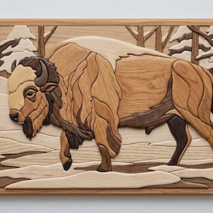 May include: Wooden wall art showing a bison in a snowy forest. The artwork is made from different wood tones, forming a detailed mosaic. The bison is brown and tan, with a light head and a dark brown beard. The frame is a light brown.