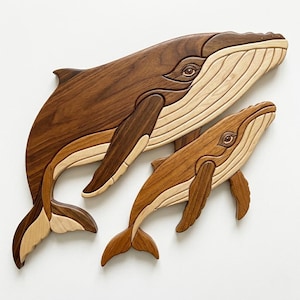 Peut inclure: Deux œuvres d'art murales en bois représentant des baleines. La plus grande baleine est brun foncé, avec des accents brun clair et beige. La plus petite baleine est brun clair. Les deux ont des yeux et des nageoires détaillés.