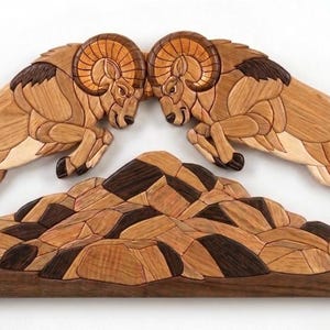 May include: Wooden wall art depicting two bighorn sheep clashing horns above a mountain range. The artwork is crafted from various wood tones, including light and dark browns, creating a detailed mosaic effect. The base is a dark brown rectangle.