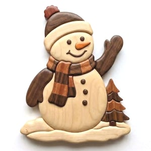 May include: A handcrafted wooden snowman wall art piece featuring a light-coloured body, a dark brown hat with a pom-pom, a striped scarf, and three circular buttons. The snowman stands on a light wood base next to a small dark wood evergreen tree.
