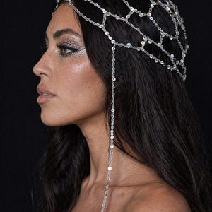 May include: A silver headpiece with a net-like design, adorned with small, reflective sequins. A strand of sequins dangles down. The headpiece is worn over dark hair against a black background. The image shows a side profile.