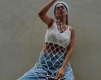 May include: A white, open-weave net top over a white crop top and light blue jeans. The person is wearing a light blue beanie, glasses, and holding a coffee cup. The outfit is set against a neutral-colored wall.