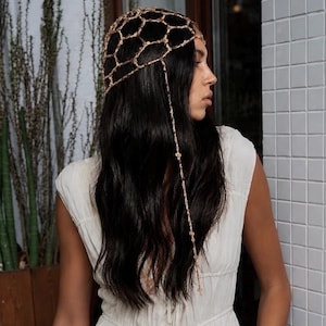 May include: A woman wearing a gold-colored headpiece with a geometric design. The headpiece is made of interconnected shapes and hangs over dark hair. She is wearing a white dress.