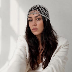 Può includere: Un copricapo argentato con un delicato design a rete, indossato sulla testa. Il copricapo è fatto di piccole perline o maglie interconnesse. La persona indossa un blazer bianco. I capelli sono lunghi e scuri.