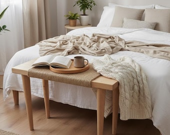 May include: A light wood bench with a woven jute seat top, positioned at the foot of a bed. The bench holds a round wooden tray with an open book and a ceramic mug, alongside a draped cream-colored cable knit throw blanket.