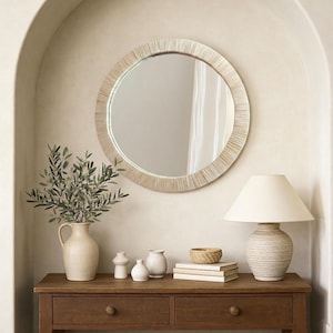 May include: A round wall mirror with a textured, light-colored frame featuring fine vertical lines. The mirror is positioned above a wooden console table holding a ceramic pitcher with olive branches, three small vases, a woven bowl, and a lamp.