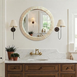 May include: A round wall mirror featuring a textured, light-colored frame with a radial pattern. The mirror is mounted on a wall above a marble countertop with a brass faucet, flanked by two wall-mounted lamps with pleated, off-white shades.