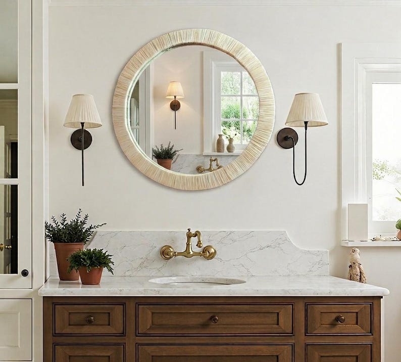 May include: A round wall mirror featuring a textured, light-colored frame with a radial pattern. The mirror is mounted on a wall above a marble countertop with a brass faucet, flanked by two wall-mounted lamps with pleated, off-white shades.