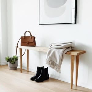 May include: A light-toned wooden bench with four tapered legs, featuring a brown leather handbag, a folded beige throw blanket with fringe, and a pair of black ankle boots placed on the floor. A gray ribbed planter with a green succulent sits nearby.