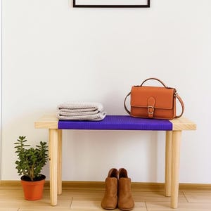 May include: A wooden bench with a purple woven seat, holding a tan satchel, folded blanket, and brown ankle boots. A small potted plant sits to the left. The bench has light-colored wooden legs.