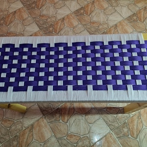 May include: A rectangular bench with a woven top in a pattern of white and purple. The bench has a wooden frame with light-colored legs. The woven top is made of thick, durable material.