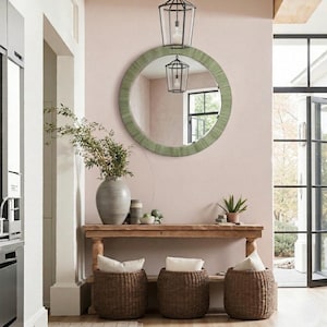 May include: A round mirror with a textured sage green frame hangs above a wooden console table. On the table sits a large gray ceramic vase with green branches, a small potted plant, and three woven baskets with white cushions underneath.