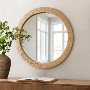 May include: A round wall mirror featuring a wide frame wrapped in light tan natural fibers. The mirror reflects a bright room with white window frames. The design is circular with a textured, linear pattern created by the tightly wound material.