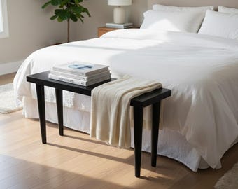 May include: A black bench with angled legs holds a stack of books and a folded cream-colored blanket. The bench is positioned at the foot of a bed with white bedding. The room has a neutral color palette with a plant in the background.