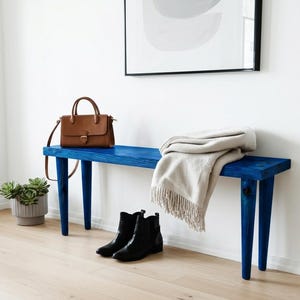 May include: A rectangular wooden bench finished in a vibrant blue stain with four tapered legs. A tan leather handbag sits on the left side, while a folded beige fringed throw blanket rests on the right. A pair of black ankle boots sits below.