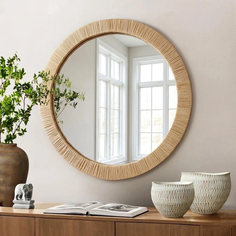 May include: A large round wall mirror featuring a frame wrapped in light tan natural fibers. The mirror reflects a bright room with white window frames. The design is circular with a textured, fibrous border surrounding the reflective glass surface.