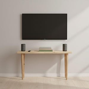 Puede incluir: Una moderna configuración de sala de estar con un televisor negro grande montado en una pared gris claro. Una consola de madera debajo sostiene dos altavoces cilíndricos, libros y un mando a distancia. La habitación tiene suelo de madera clara.