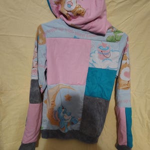 Puede incluir: Una sudadera con capucha de patchwork con una capucha rosa con un gráfico de oso. El cuerpo de la sudadera con capucha es un patchwork de paneles rosas, azules y grises con gráficos de Care Bears. Las mangas son una mezcla de rosa y gris.
