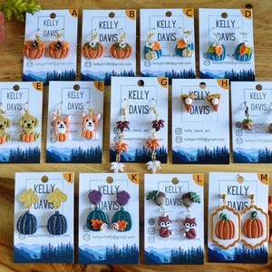 May include: A collection of thirteen pairs of handmade polymer clay earrings displayed on branded cards labeled A through M. The designs feature seasonal autumn themes including pumpkins, foxes, dogs, acorns, leaves, and pie slices with metal hooks.