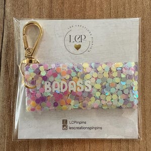 May include: A rectangular keychain with the word "BADASS" in white letters, filled with colorful confetti. It has a gold-colored clasp and is packaged in a clear plastic sleeve. The keychain is on a light-colored background.