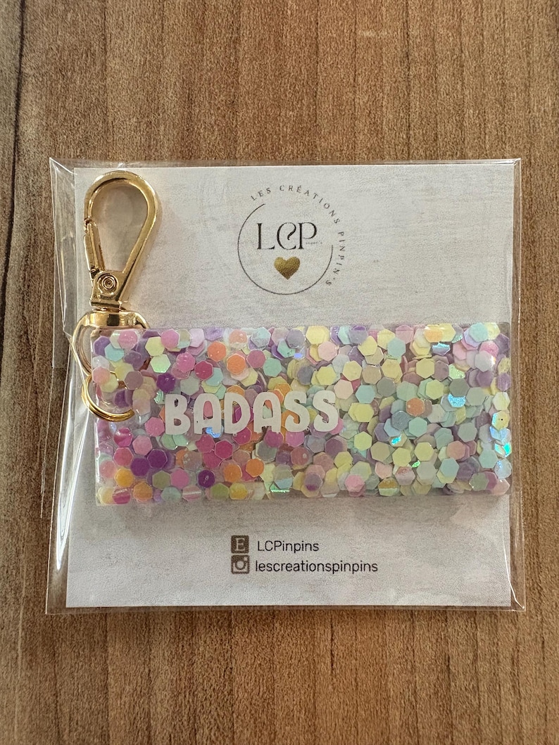 May include: A rectangular keychain with the word "BADASS" in white letters, filled with colorful confetti. It has a gold-colored clasp and is packaged in a clear plastic sleeve. The keychain is on a light-colored background.
