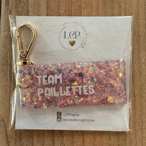 May include: A rectangular keychain with pink glitter and the words "TEAM PAILLETTES" in white. It has a gold-colored clasp and is packaged in clear plastic. The keychain is on a white card with a logo and social media handles.