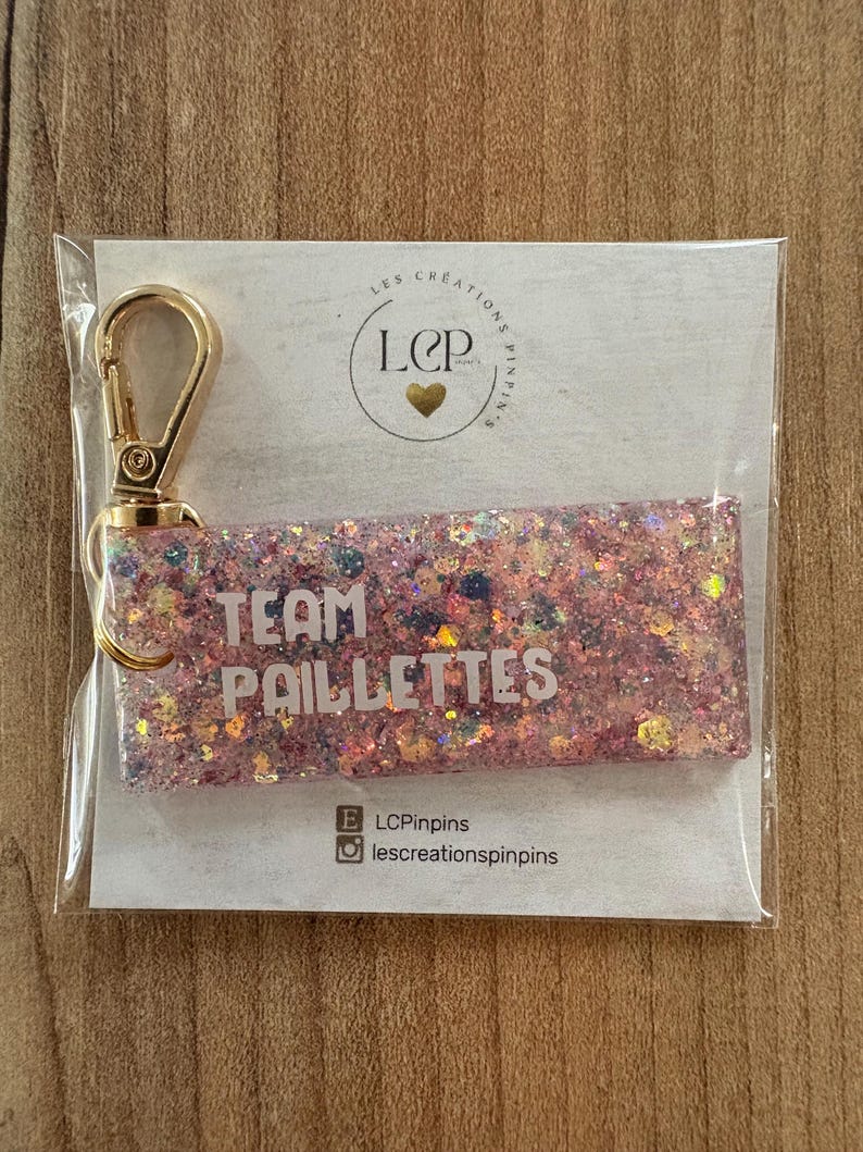 May include: A rectangular keychain with pink glitter and the words "TEAM PAILLETTES" in white. It has a gold-colored clasp and is packaged in clear plastic. The keychain is on a white card with a logo and social media handles.