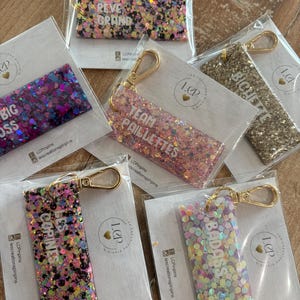 Puede incluir: Una colección de llaveros con purpurina en varios colores, cada uno con un cierre dorado. Los llaveros presentan frases como "REVE GRAND", "BIG BOSS", "TEAM PAILLETTES", "LES COPINES", "BICHETTE" y "BADASS".