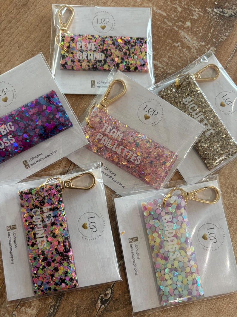 May include: A collection of glitter keychains in various colors, each with a gold-colored clasp. The keychains feature phrases like "REVE GRAND", "BIG BOSS", "TEAM PAILLETTES", "LES COPINES", "BICHETTE", and "BADASS".