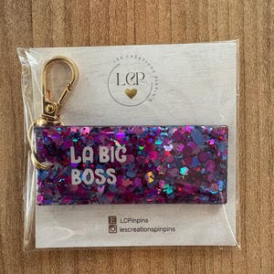 May include: A rectangular keychain with the text "LA BIG BOSS" in white letters, set against a background of purple and blue glitter. The keychain has a gold-colored clasp and is packaged in a clear plastic sleeve.