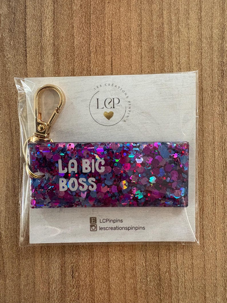 May include: A rectangular keychain with the text "LA BIG BOSS" in white letters, set against a background of purple and blue glitter. The keychain has a gold-colored clasp and is packaged in a clear plastic sleeve.