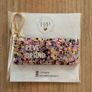 May include: A rectangular keychain with a gold-colored clasp. The keychain is filled with colorful glitter and the words "REVE GRAND" in white. The keychain is packaged on a white card with a logo and social media handles.