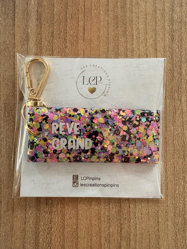 May include: A rectangular keychain with a gold-colored clasp. The keychain is filled with colorful glitter and the words "REVE GRAND" in white. The keychain is packaged on a white card with a logo and social media handles.