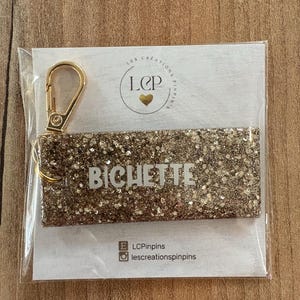 May include: A gold glitter keychain with the name "BICHETTE" in white lettering. The keychain is attached to a gold-colored clasp. The product is packaged on a white card with a logo and social media handles.