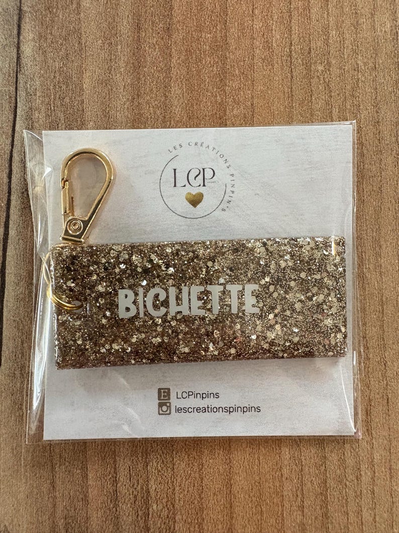 May include: A gold glitter keychain with the name "BICHETTE" in white lettering. The keychain is attached to a gold-colored clasp. The product is packaged on a white card with a logo and social media handles.