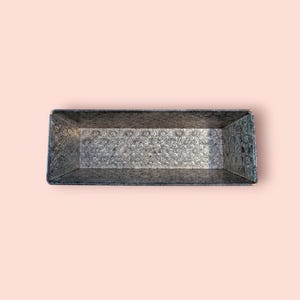 May include: A rectangular, galvanised metal tray with a floral pattern. The tray has a slightly distressed appearance, with visible texture and a dark grey colour. The sides are angled, and the tray is empty, set against a light pink background.