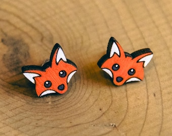 Könnte beinhalten: Ein Paar hölzerne Fuchsohrringe mit orangefarbenem Fell und schwarzen Augen. Die Ohrringe liegen auf einer Holzoberfläche.