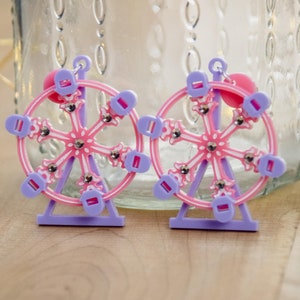 May include: A pair of purple and pink earrings shaped like a Ferris wheel. The earrings have a pink wheel with white spokes and purple accents.