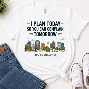 May include: White t-shirt with the text "I PLAN TODAY - SO YOU CAN COMPLAIN = TOMORROW" in green and black. A colorful city skyline graphic is below the text. The shirt is paired with blue jeans and white sneakers.