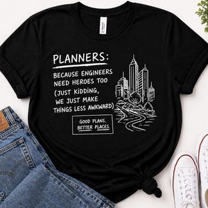 May include: Black t-shirt with white text and a cityscape graphic. The text reads "PLANNERS: BECAUSE ENGINEERS NEED HEROES TOO (JUST KIDDING, WE JUST MAKE THINGS LESS AWKWARD) GOOD PLANS. BETTER PLACES."