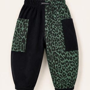 May include: Corduroy pants featuring a split design with one solid black leg and one green and black leopard print leg. The pants include a black elastic waistband with a ZIGGYLOVESMICKY label, two black pockets on the leopard side, and two leopard pockets on the black side.