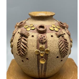 May include: A beige ceramic vase with a rounded body and a narrow neck. The vase is decorated with gold-coloured accents, including circular and leaf-like designs, and vertical stripes. The design includes small, evenly spaced holes.