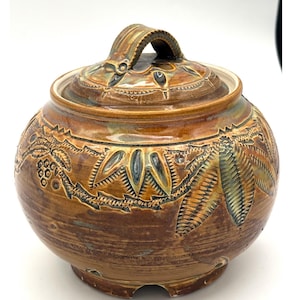 Op de afbeelding: Een bruine keramische pot met een deksel en handvat. De pot heeft ingewikkelde gesneden ontwerpen van bladeren en andere patronen. Het kleurenpalet omvat bruintinten, beige en hints van groen en blauw. De pot is rond met een voet.