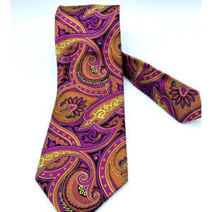 Puede incluir: Una corbata estampada con un diseño de cachemira en tonos morado, naranja y amarillo. La corbata está hecha de una tela suave y se muestra sobre un fondo blanco. El diseño incluye patrones arremolinados y florales.