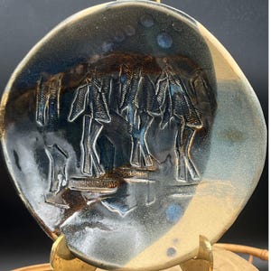 May include: A handcrafted ceramic plate with a unique design. The plate features a dark blue and brown glaze with an embossed image of figures. The plate is displayed on a gold-coloured stand.
