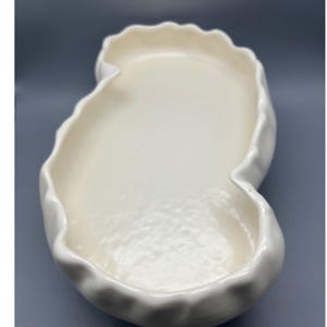 Puede incluir: Un plato de cerámica blanco en forma de riñón con un borde con volantes. El plato tiene un interior liso y brillante y un borde ondulado y texturizado. El diseño es simple y elegante, adecuado para servir o exhibir.