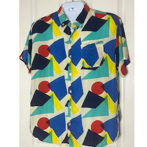 May include: Short-sleeved button-down shirt with a colorful geometric pattern. The shirt features a cream-colored background with shapes in red, blue, yellow, and teal. It has a chest pocket and a button-down collar.