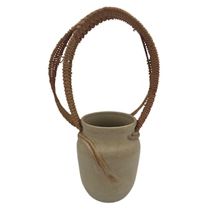 Peut inclure: Un vase en céramique beige avec une anse marron tressée. Le vase a un corps arrondi et une surface texturée. Une bande décorative marron entoure le vase. La poignée est fixée au vase par un nœud.